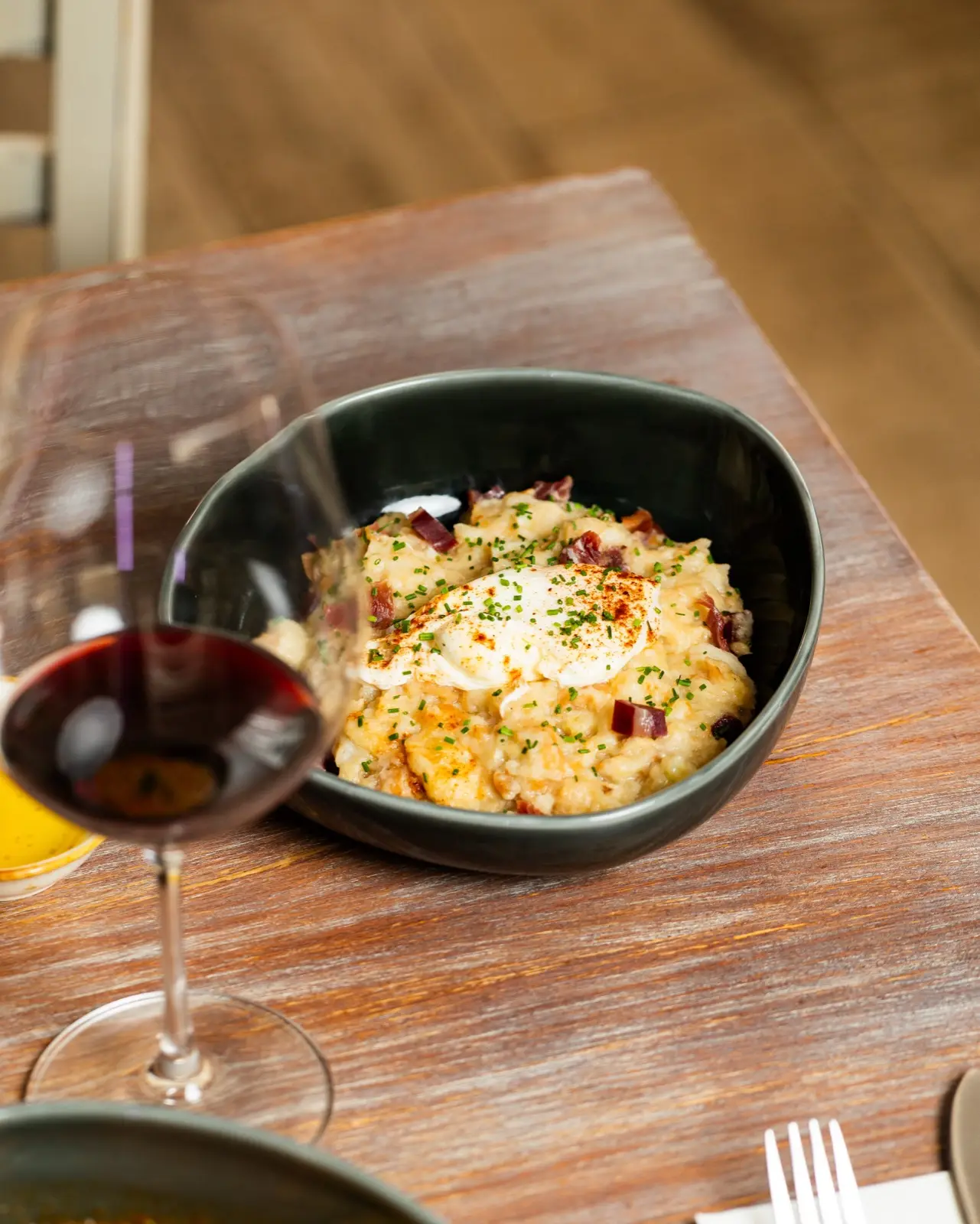Huevos rotos con jamón en un cuenco oscuro, junto a una copa de vino.