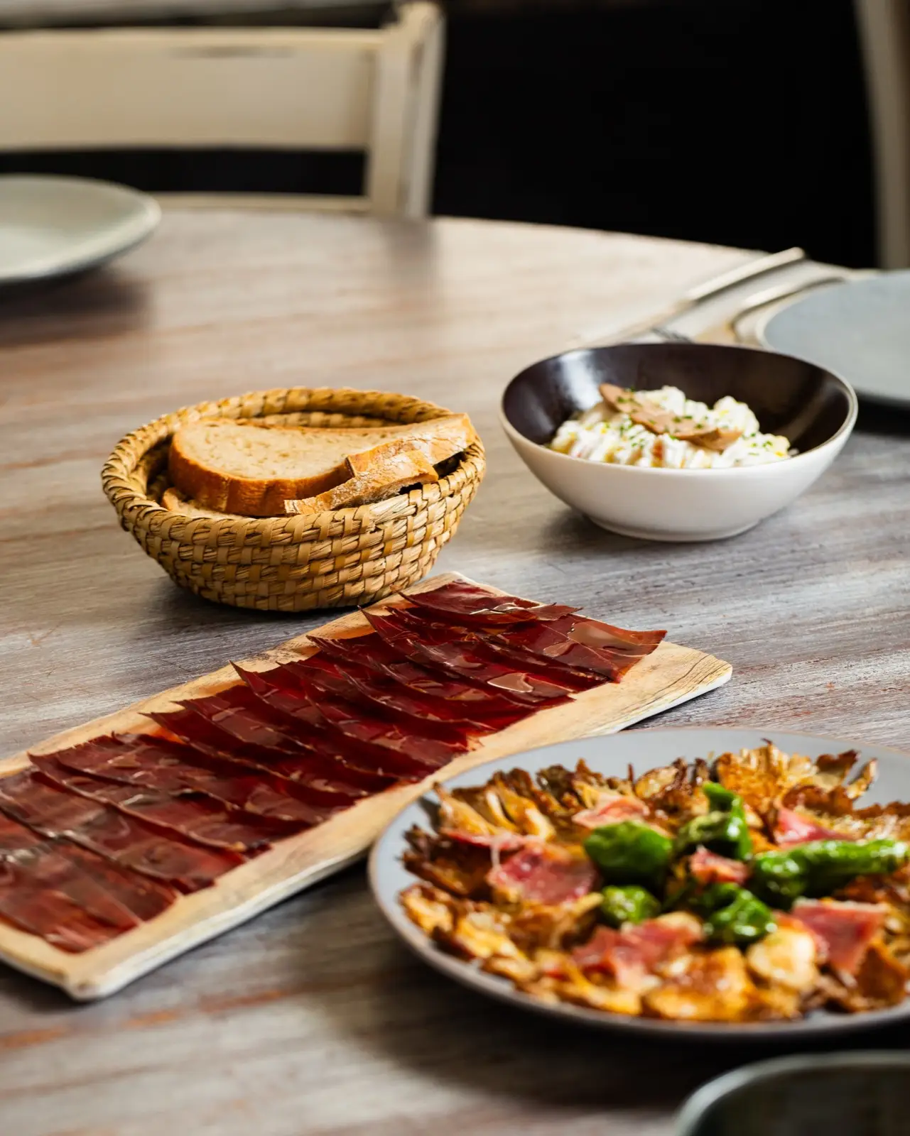 Lonchas de jamón sobre una tabla de madera junto a una cesta de pan, con setas y ensaladilla al fondo.