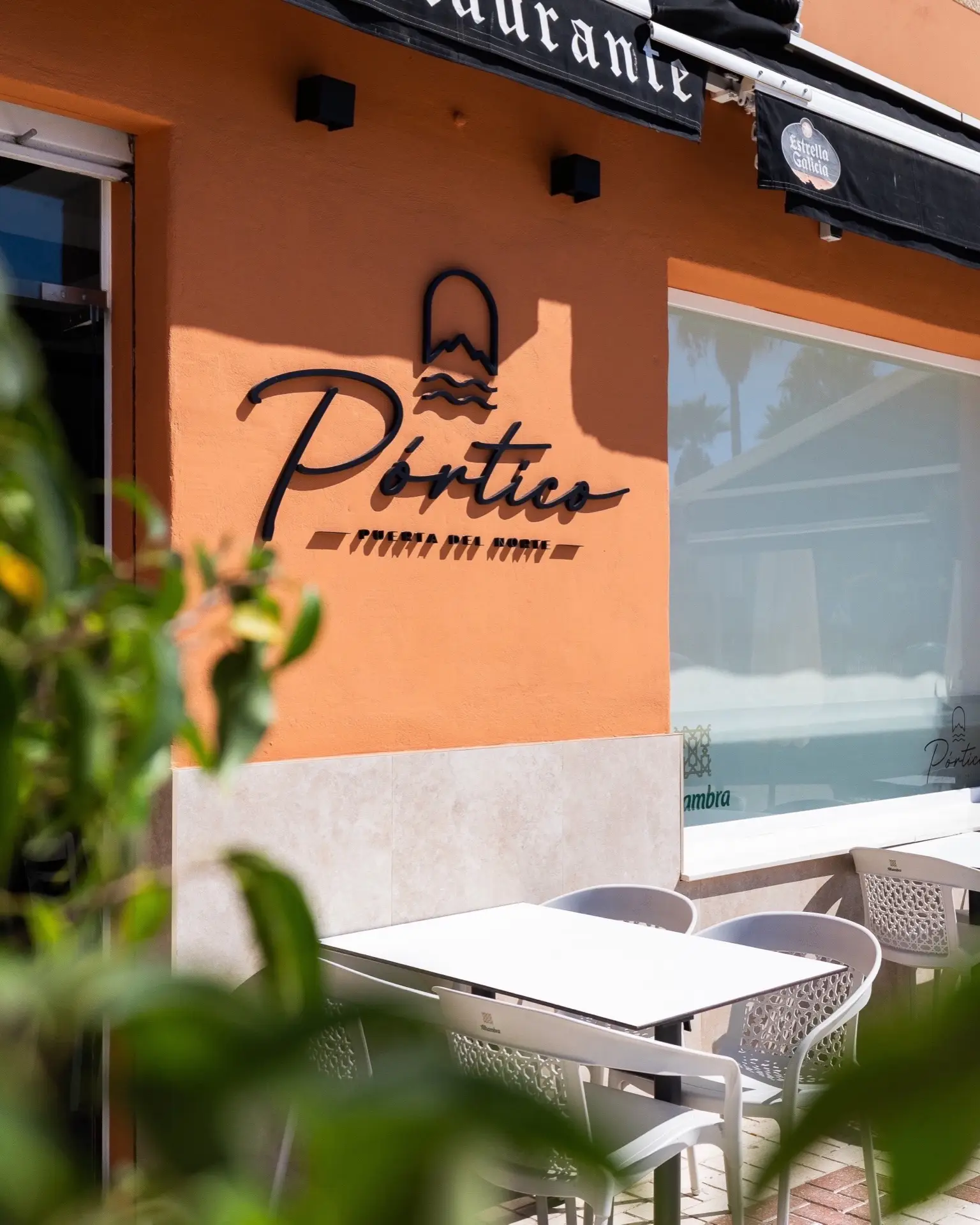 Fachada terracota del restaurante Pórtico con su logotipo, ventanal y terraza con mesas y sillas blancas, vistas entre hojas.