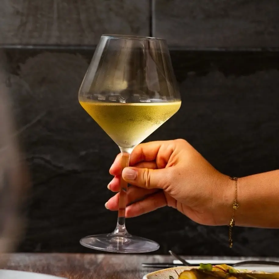 Una mano sostiene una copa de vino blanco frente a una pared de pizarra oscura, con platos en primer plano.