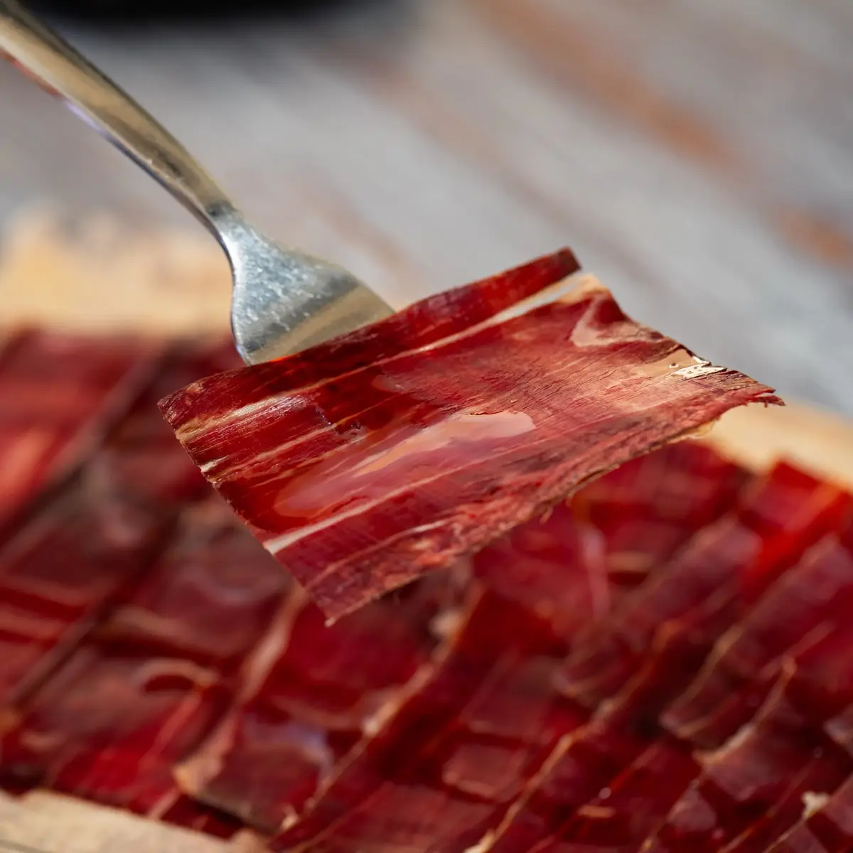 Primer plano de una loncha de jamón levantada con un tenedor sobre más lonchas en una tabla de madera.