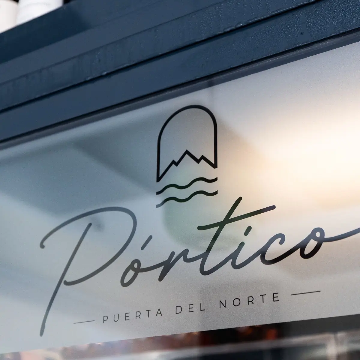 Logotipo de Pórtico — Puerta del Norte vinilado en el escaparate del restaurante, con botellas de vino en la balda de arriba.