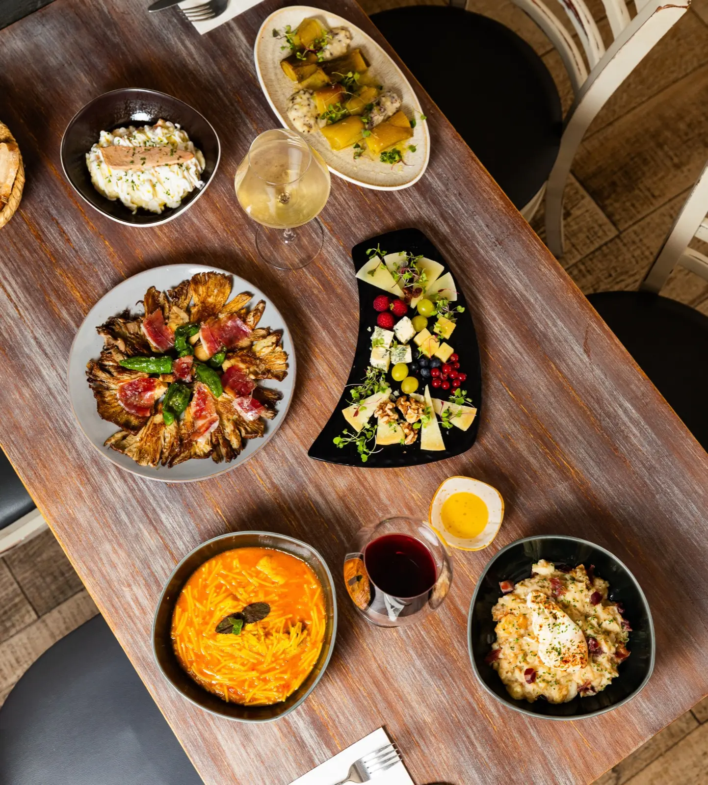 Mesa vista desde arriba con platos de setas con jamón, puerro confitado, tabla de quesos, huevos rotos y salmorejo, junto a copas de vino.
