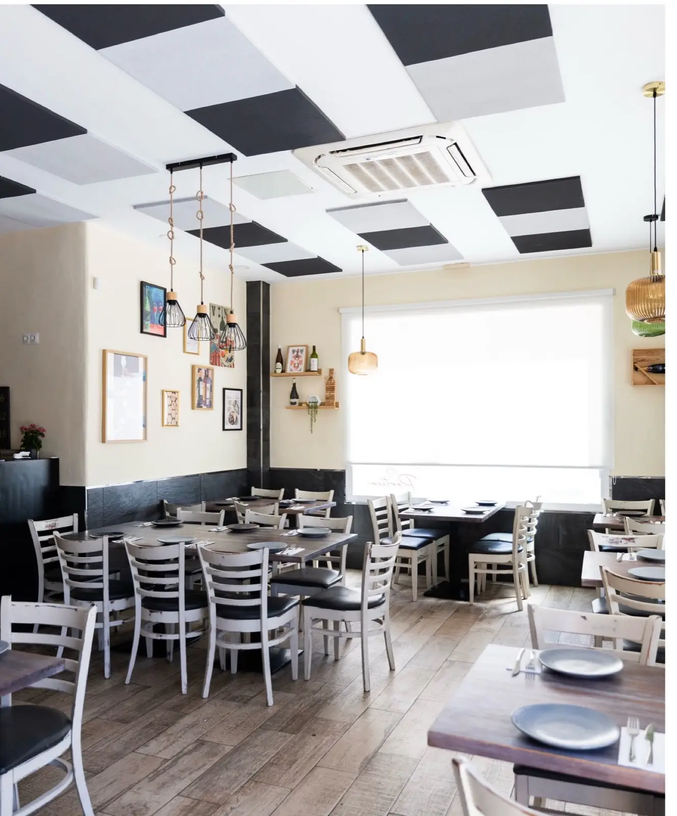 Sala del restaurante con techo de damero blanco y negro, sillas blancas, mesas de madera y una gran ventana con luz natural.